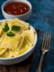 In the photo we see a portion of ravioli drizzled with melted butter. On top is a sprig of fresh parsley. There is a fork to the right of the bowl. In the background, we see sauce in a bowl...