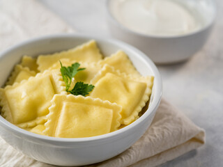 In the photo we see ravioli in a deep white bowl. In the upper right corner of the photo is a white bowl of sour cream. The ravioli is covered with a piece of green herbs. White napkin..