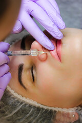 Obraz na płótnie Canvas A cosmetologist injects a hyaluronic acid-based filler into the patient's lips. The process of lip augmentation in a young beautiful woman. A nurse is wearing purple rubber gloves. Beauty injections.