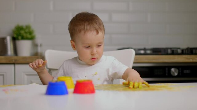 Handsome little baby boy draws with his hand on the kitchen table with finger paints. Slow motion