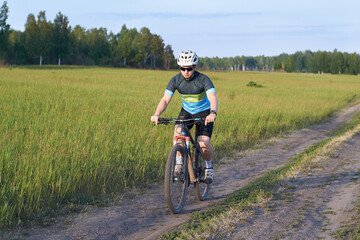 Naklejka premium The guy is a cyclist leisurely rides a mountain bike on a dirt road. Dressed in a bicycle uniform, helmet and sunglasses. Endurance training. Healthy lifestyle and weight loss. Sunny weather.