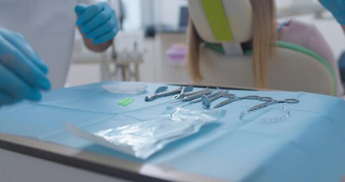 Dental scissors, dental probes, scaler, mirror, braces archwire, tweezers are laying on the table and a medical worker is taking out dental forceps of a sterile bag and putting them on the table.