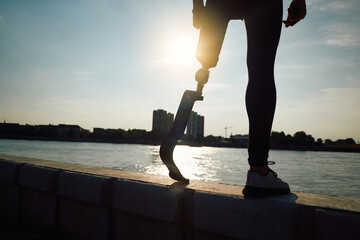 Unrecognizable handicapped sportsman with artificial leg standing on riverbank at sunset.