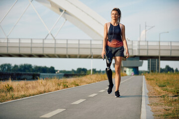 Young smiling sportswoman with leg prosthesis walks on a path outdoors and looking at distance.