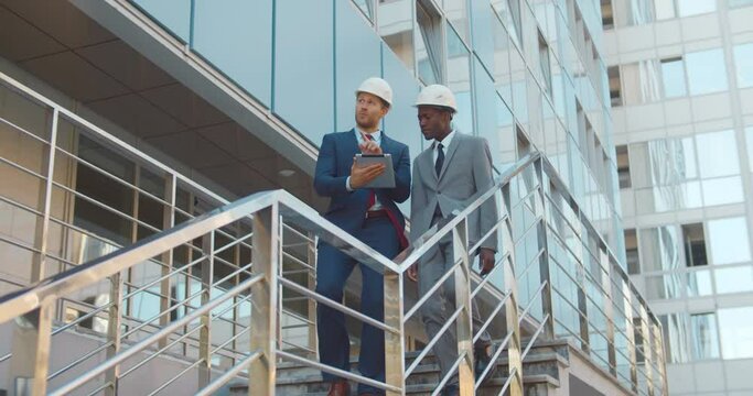 Diverse business partners in hardhat walking downstairs outside modern building and using digital tablet