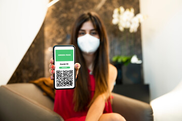 Defocused young girl wearing a face mask, holding a smartphone with a Green Pass. The Green Pass...