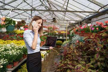 Smiling Greenhouse owner posing with a laptop in her hands talking on the phone having many flowers in background and glass roof. Space for text on screen.