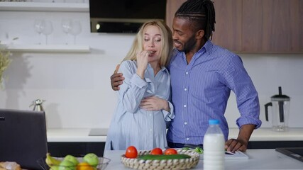 Pregnant Caucasian woman tasting organic ingredients for healthful salad as African American man hugging spouse talking in slow motion. Portrait of interracial couple cooking breakfast at home - Powered by Adobe