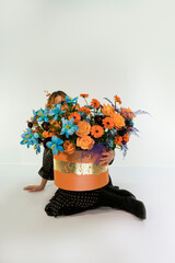 A girl in black dress sitting on white background with orange hat box with floral arrangement. Blue burgundy orange color flowers.