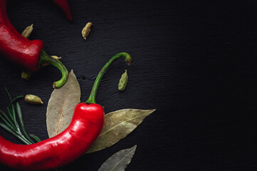 Two hot red chilly pepper with laurel leaf and cardamom laying on black painted wood board, with space to add text.