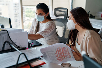 two young latina business women talking and working together in the office, wearing covid-19 protective masks in times of pandemic, teamwork concept