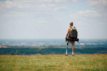 Rear view of disabled man with leg prosthesis enjoys the view from top of the hill while hiking in...