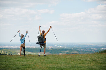 Rear view of carefree hikers with leg disability standing on top the hill with raised arms.