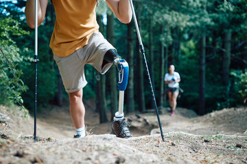 Unrecognizable hiker with prosthetic leg uses support sticks while walking through the woods.