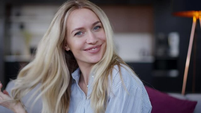 Close-up portrait of charming blond Caucasian young woman with blue eyes looking at camera smiling touching hair in slow motion. Positive happy relaxed millennial posing indoors at home in living room