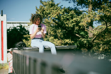 Cute young woman sitting outdoors with headphones on and choosing good music on her phone.