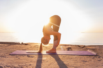 Young blonde woman in sportswear doing yoga asanas Uttanasana pose on the seashore at sunrise on purple yoga mat. Sports meditation by the ocean on the yellow sand. Positive effects on brain function