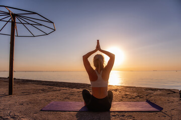 Young blonde woman in sportswear meditates yoga asanas Sukhasana Pose on the seashore at sunrise on...