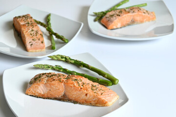 Close up of white plates with grilled salmon fillets with asparagus on white surface, with copy space