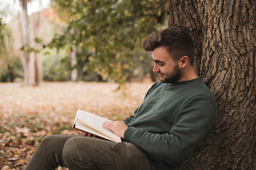 Young Caucasian man sitting next to the tree and reading a book