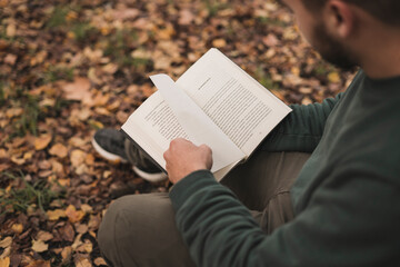 Young Caucasian man sitting next to the tree and reading a book