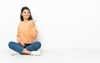 Teenager Russian girl sitting on the floor pointing to the side to present a product