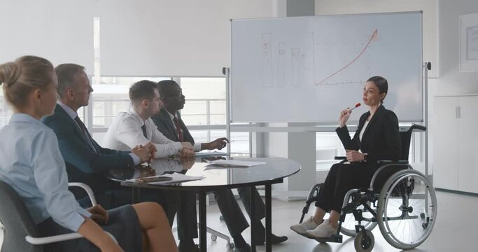 Young woman in wheelchair teaching group of business people at whiteboard in office