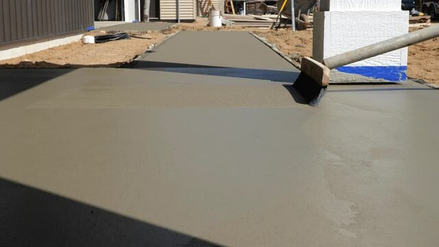 A wide push broom is pulled lightly over the top of a new cement concrete sidewalk, to finish it and leave a slightly rough walking surface. Closeup clip of three strokes as another worker approaches.
