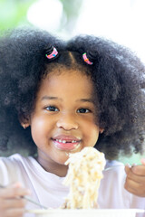 A little curly-haired African American girl sits at the table eating delicious Spaghetti Carbonara. Fun, cheerful. Appetite. Childhood and eating concepts.