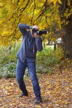 Male photographer with a camera, taking pictures in autumn, among fallen leaves and yellow trees, front view
