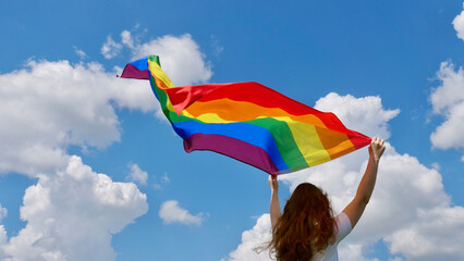 Blonde, lesbian, woman holding a rainbow LGBT gender identity flag on sky background with clouds on...