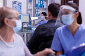 Young nurse wearing protection mask and visor against coronavirus infection typing on computer new patients appointments. People sitting in crowned reception room waiting for doctor consultation.