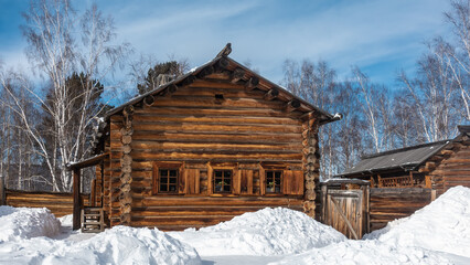 An old wooden house was built from natural unpainted logs. There are shutters on the windows. Plank fence. There are snowdrifts on the ground. Bare birch trees against the blue sky. Siberia