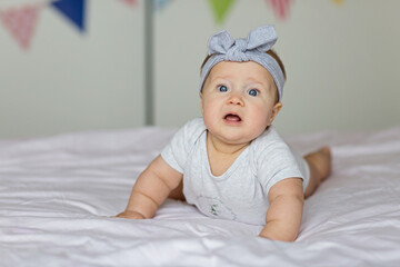 Caucasian blonde baby six months old lying on bed at home. Kid wearing cute gray clothing. Bedroom decorated with flags.
