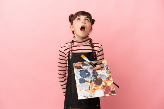 Little artist girl holding a palette isolated on pink background looking up and with surprised expression