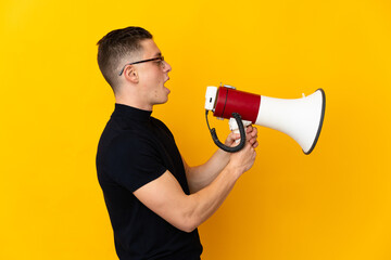 Young caucasian man isolated on yellow background shouting through a megaphone to announce something in lateral position