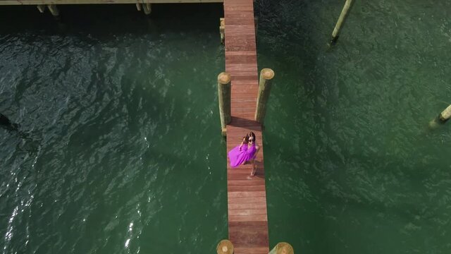 Aerial View of Lonely Young Woman in Purple Dress Standing on Wooden Dock Above Sea Water, Fashion Concept. Top Down Drone Shot