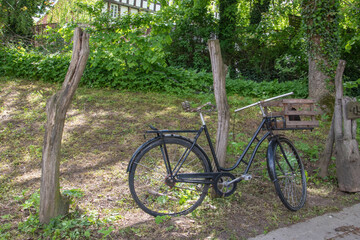 Fototapeta premium Altes schwarzes Fahrrad mit einem Holzkorb vor dem Lenker lehnt an einem Holzpfahl.