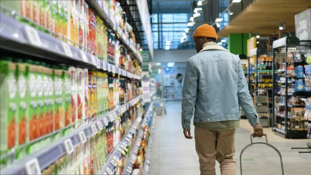 Black man shopper in jacket wanders along long grocery shop rows looking around and holding plastic trolley close backside view