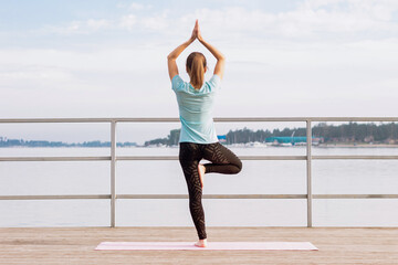 A young slim mindful woman does yoga on a mat on a pier by the sea. Peaceful girl stands on one leg in the tree pose with her folded hands up to greet the dawn. Vrikshasana asana. Morning meditation 