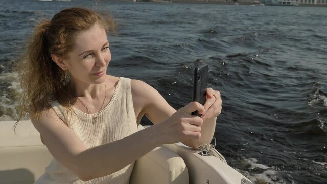 Lady with long curly hair in ponytail makes photos on black smartphone sailing along waving river water against bank close view