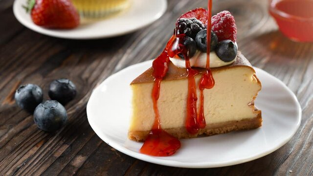 Strawberry topping pouring on fresh slice of cheesecake with blueberries, sweet and natural product, tasty baked dessert on wooden background, handmade pastry, appetizer bakery product in slow motion