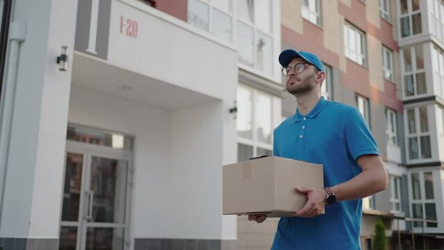 Young delivery man wearing glasses and gloves holding box use phone walking on street quarantine outdoor shopping package pandemic postman self isolation slow motion