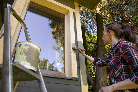 Woman worker painting wooden house exterior wall with paintbrush and wood protective color home improvement concept.