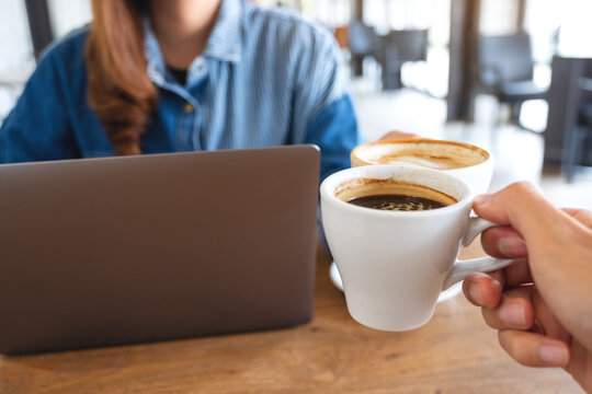 Closeup image of a woman and a man clinking coffee cups together while working on laptop computer
