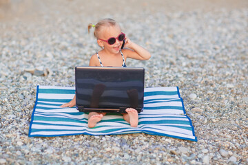 A cute little happy girl in pink sunglasses sits on a striped blue and white mat with a laptop on the beach on a warm summer day. Vacation by the sea concept. Funny kids. Sea. Ocean.