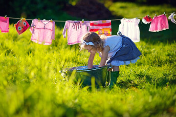 A little cute girl makes lingerie in a small aluminum pool near a clothing line in a meadow on a...