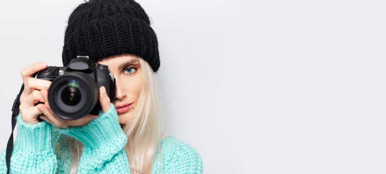 Fototapeta Portrait of blonde girl photographer taking photo on DSLR camera, on white background. Wearing black hat and blue sweater. Panoramic photo with copy space.