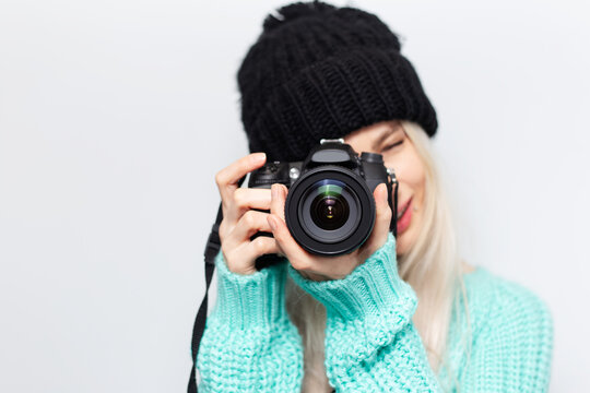 Portrait of pretty blonde girl, photographer taking photo on DSLR camera, on white background. Wearing black hat and cyan sweater.