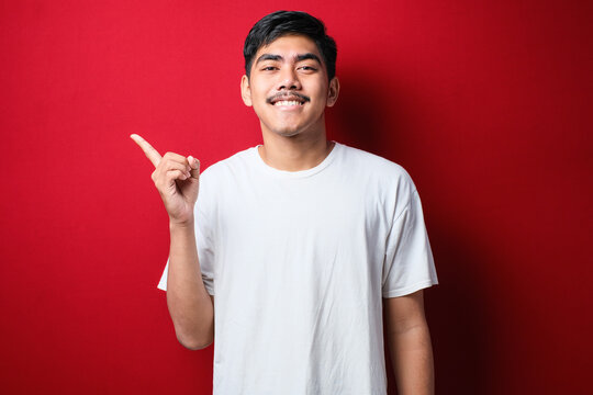 Young Handsome Asian Man Wearing White Tshirt With A Big Smile On Face; Pointing With Hand Finger To The Side Looking At The Camera.